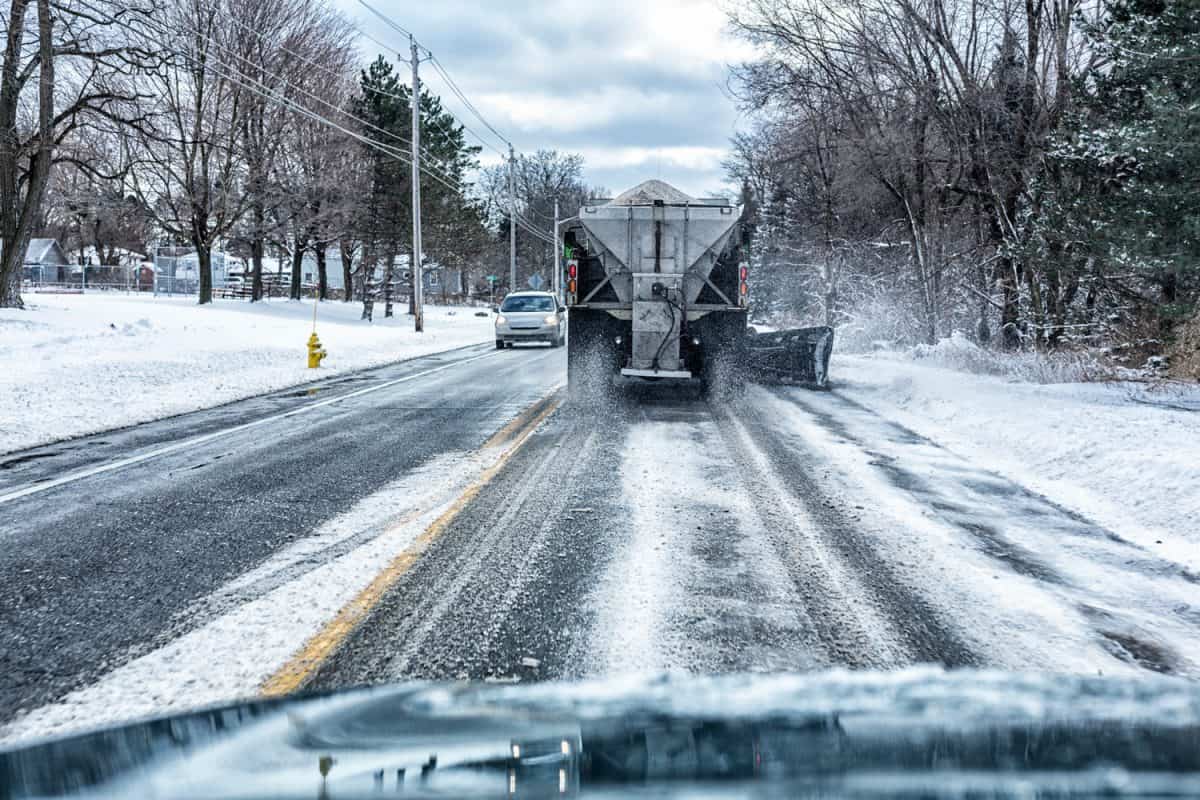 A large slow-moving snow plow dump truck is spreading rock salt on a rural highway during a snow storm. 