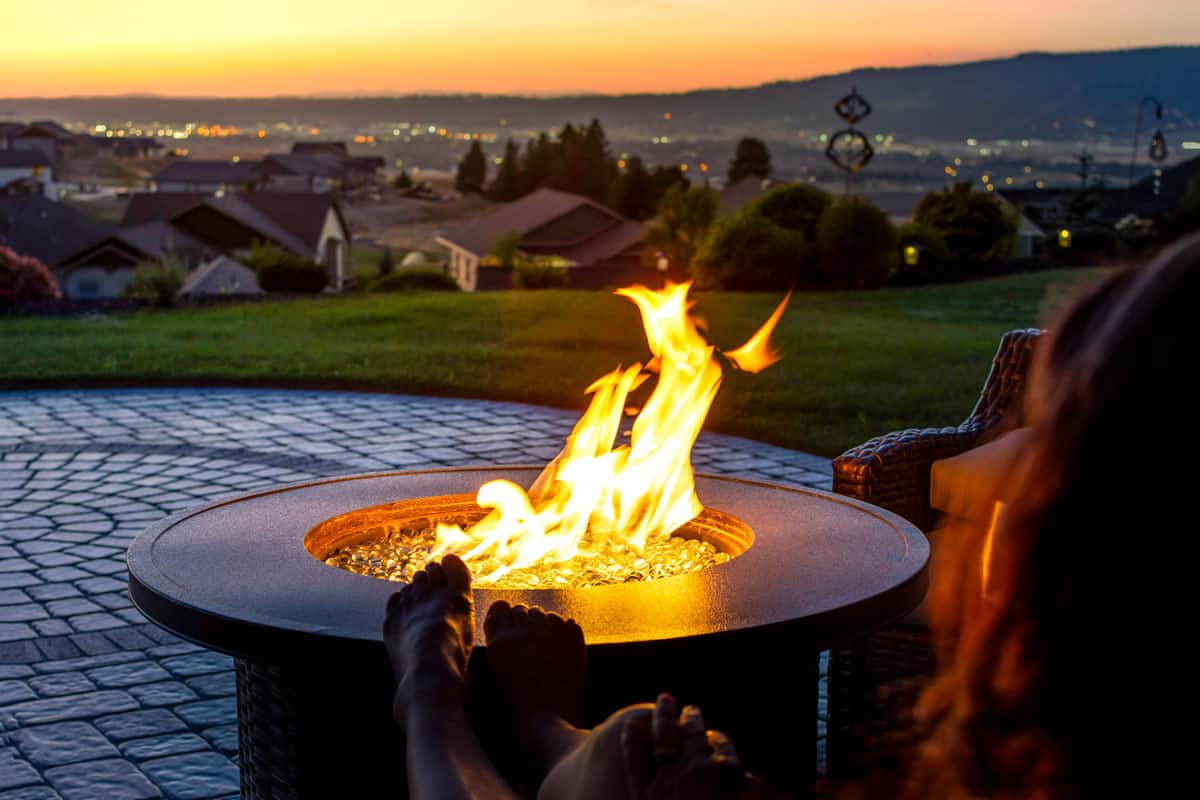 A woman relaxes by a roaring firepit on a paver patio at sunset overlooking, 15 Awesome Fire Pit Paver Ideas