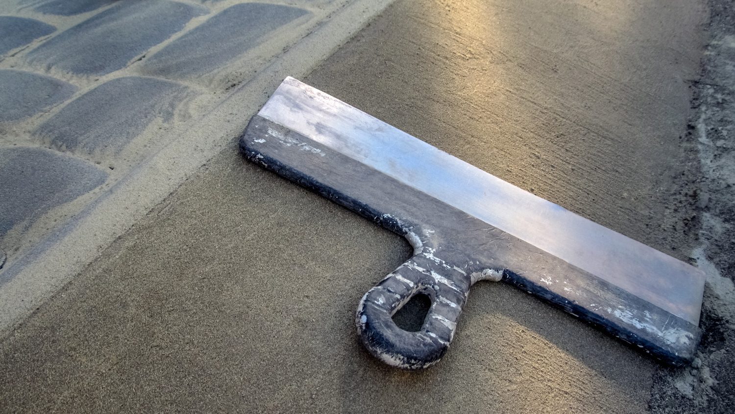 A large construction spatula lies on the surface of the concrete paver edge. 