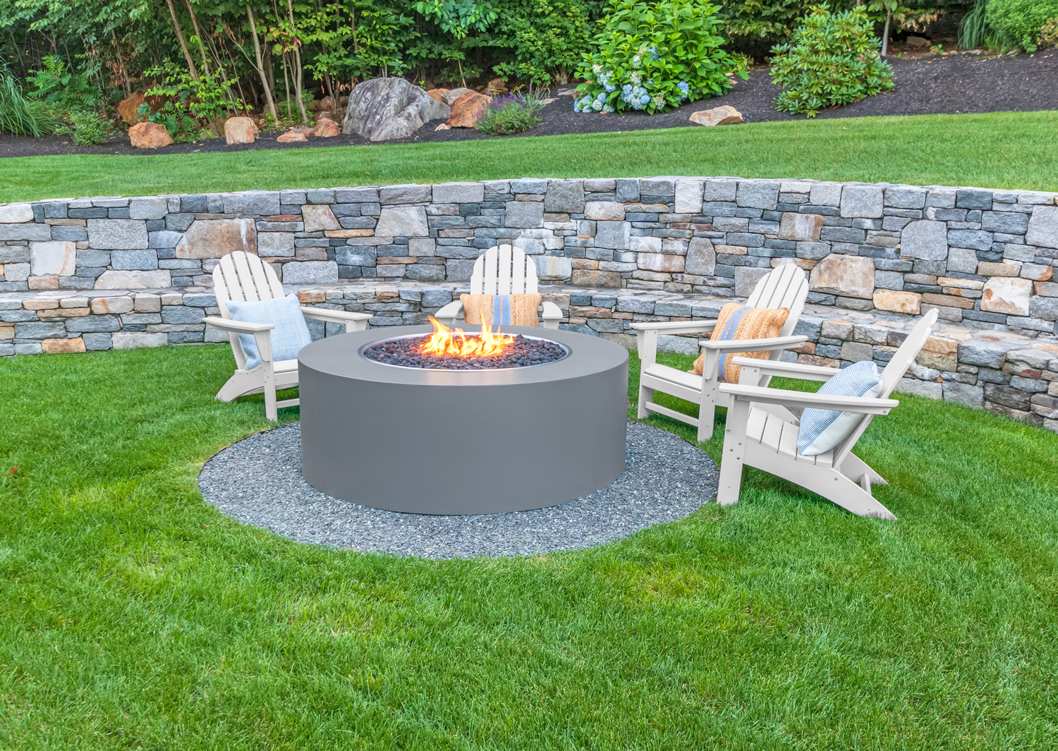 Adirondack chairs around fire pit with stone wall and lush green grass.