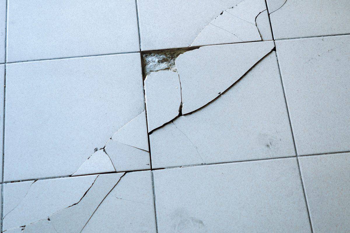 Close-up view of light gray tile with large diagonal cracks and chips.

