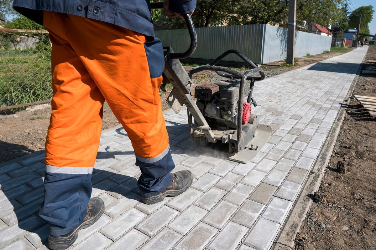 Man in orange uniform using vibrational paving stone machine for finish on a sidewalk road construction site