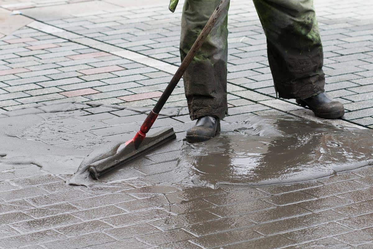 A man spreads sealer on block of paver using rubber edged spreader, Is Paver Joint Stabilizer Necessary?