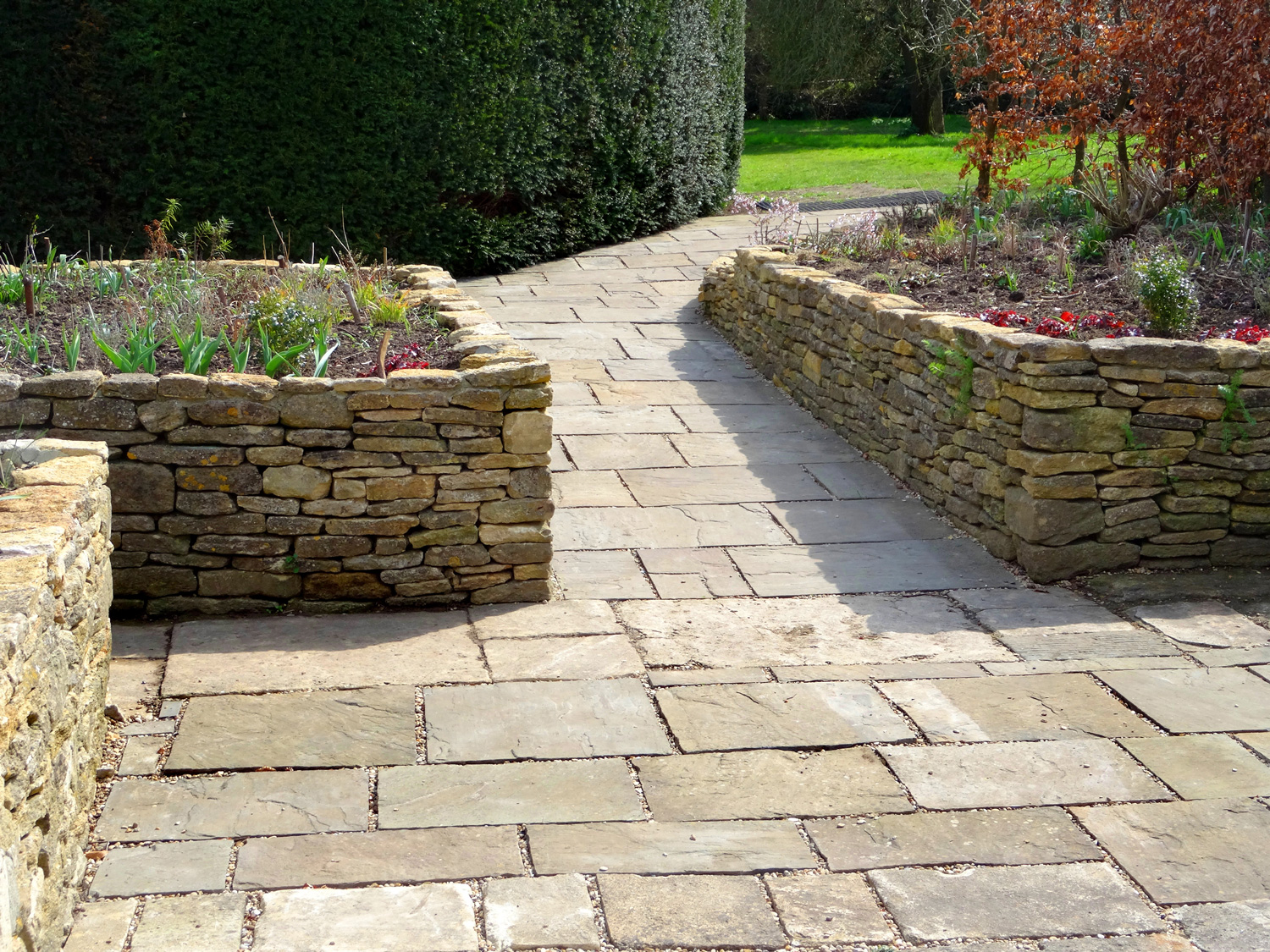 Photo showing a sunken garden with raised beds made from dry-stone walls and a patio of flagstone paving. The garden is designed to be accessible for gardeners in wheelchairs, with the sloped pathway leading to the flowerbeds.