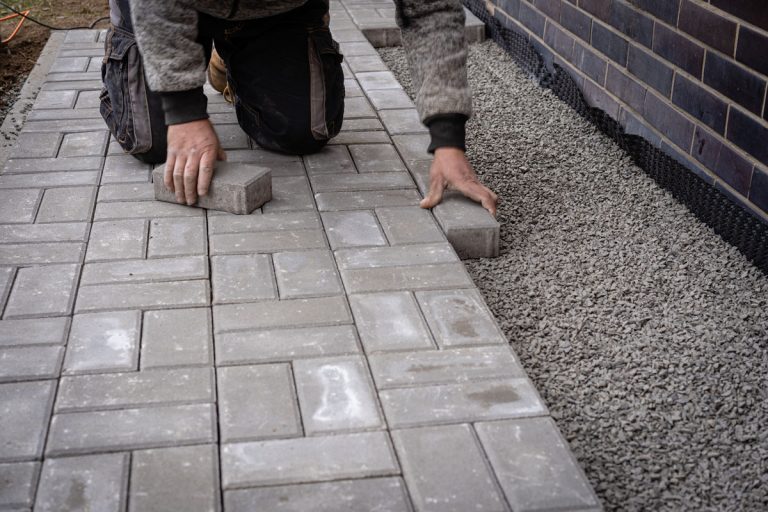 Unidentified worker is laying gray concrete paving slabs in house courtyard. Professional bricklayer is installing new tiles or slabs for walkway, sidewalk or patio on leveled gravel foundation base, Do Concrete Pavers Absorb Water?