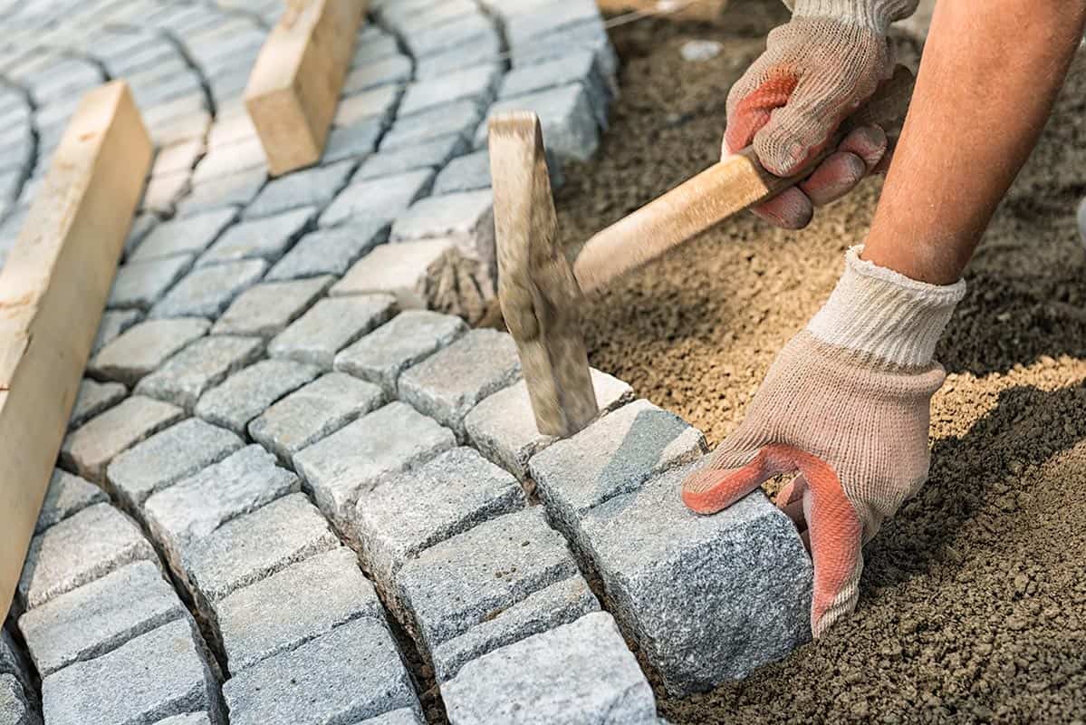 Workman's gloved hands use a hammer to place stone pavers