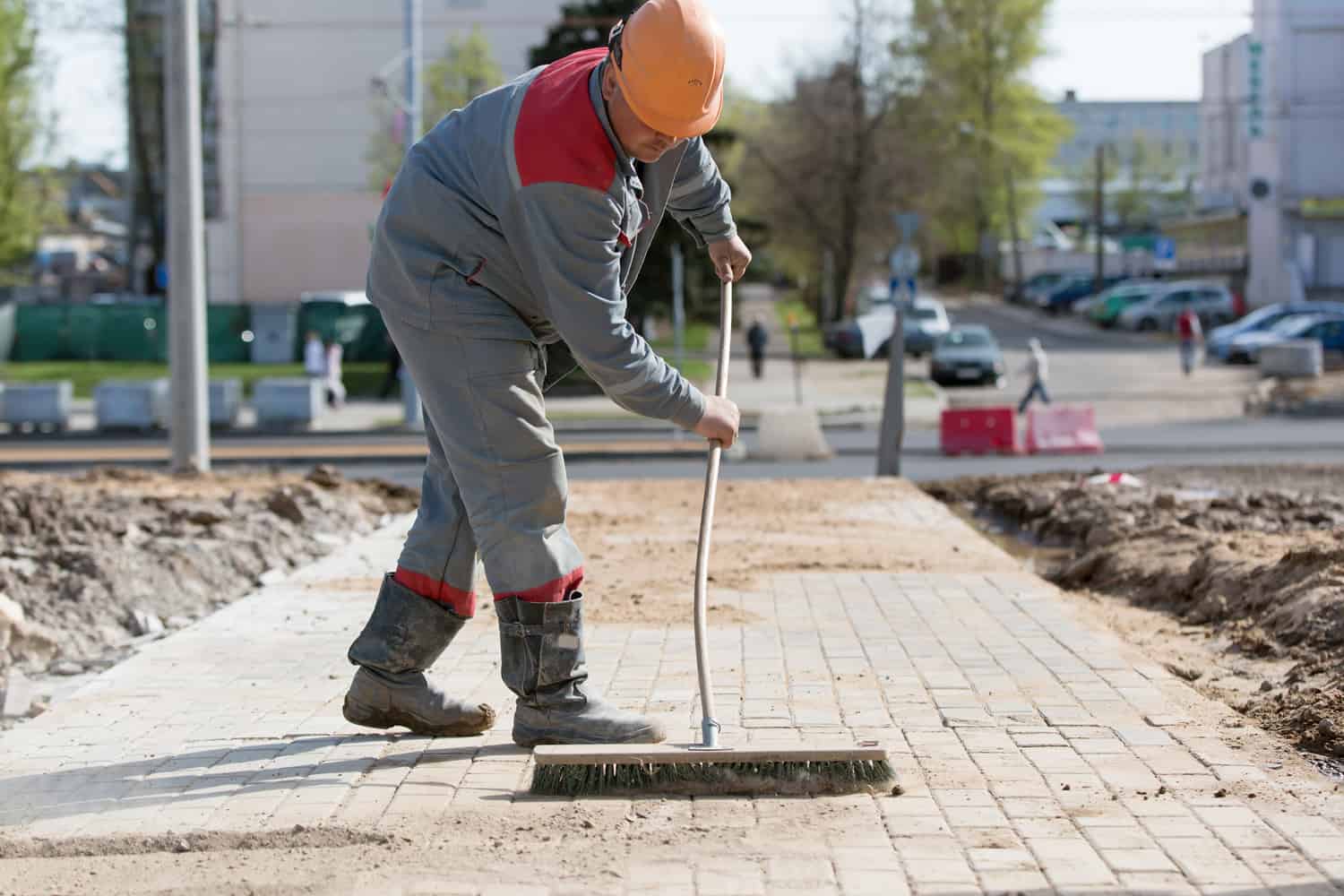 Construction worker grouting dry sand with brush into paver bricks joints during road works
